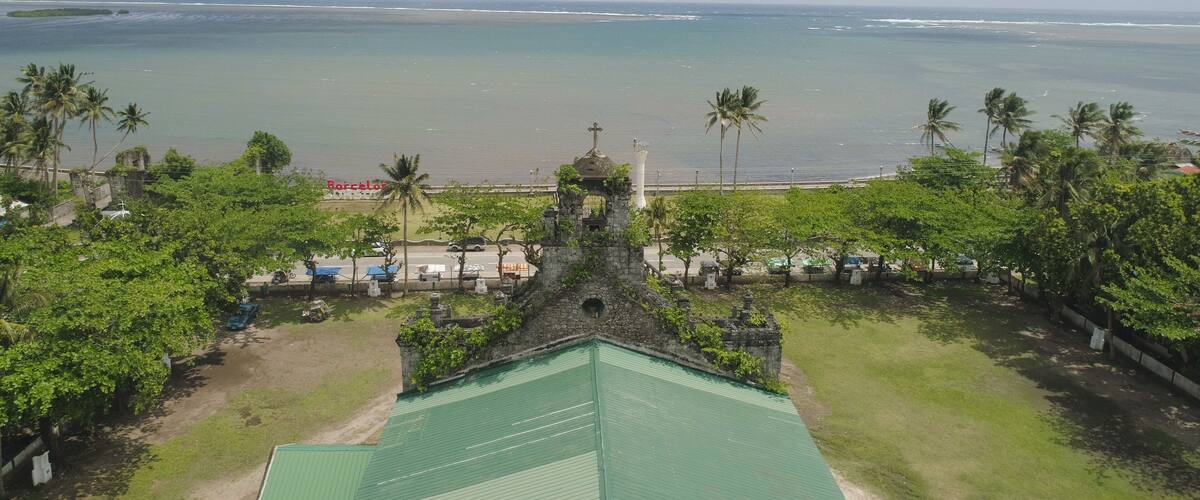 A coastal view with a beach, palm trees, and a church with a cross on top.