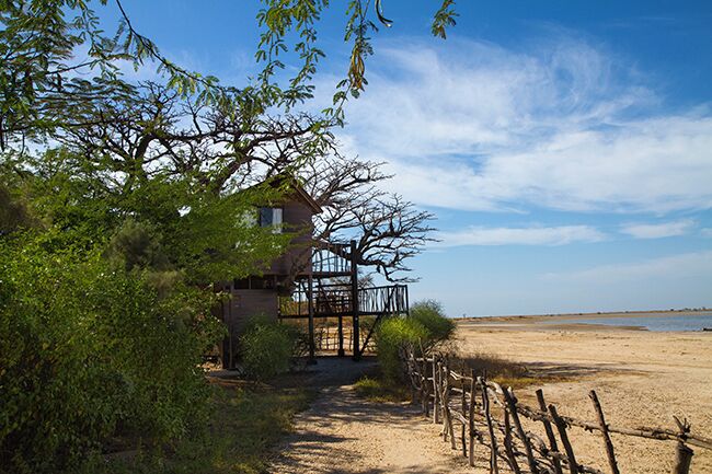 A wooden house with a balcony overlooking a sandy beach, surrounded by greenery and a clear sky.