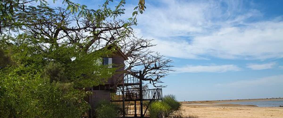 A wooden house with a balcony overlooking a sandy beach, surrounded by greenery and a clear sky.