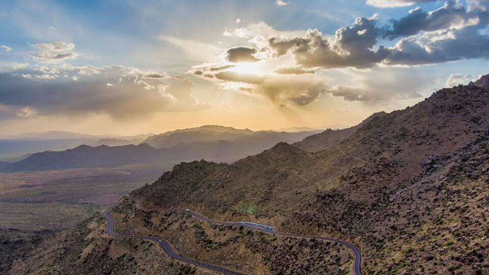 A winding mountain road with a desert landscape and distant mountains under a partly cloudy sky.