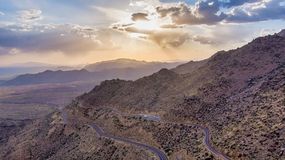 A winding mountain road in a desert landscape with distant mountains and a cloudy sky.