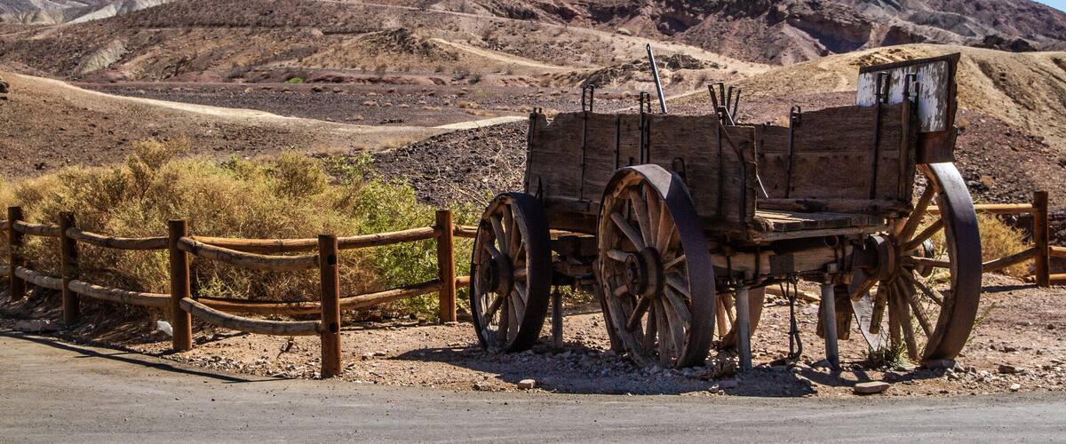 A wooden wagon with large wheels parked beside a dirt road, surrounded by a wooden fence, with mountains in the background.