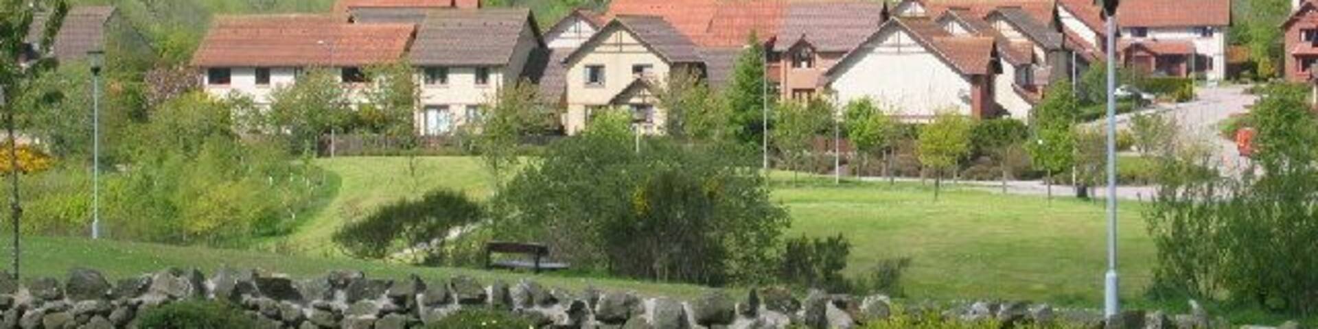 A residential area with houses, a stone wall, and a grassy hill in the background.