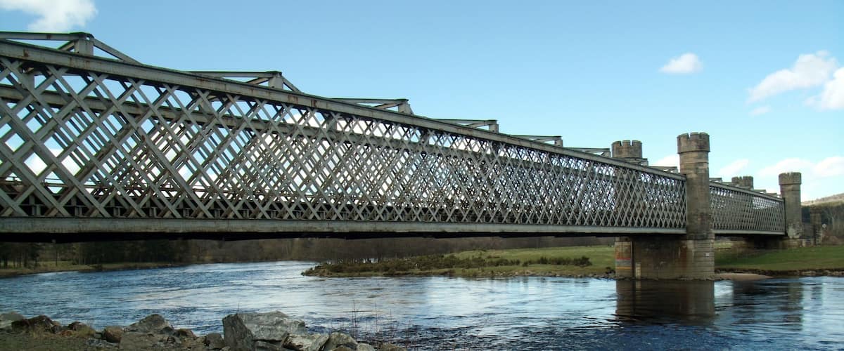 A metal bridge over a river with grassy banks and a clear sky.