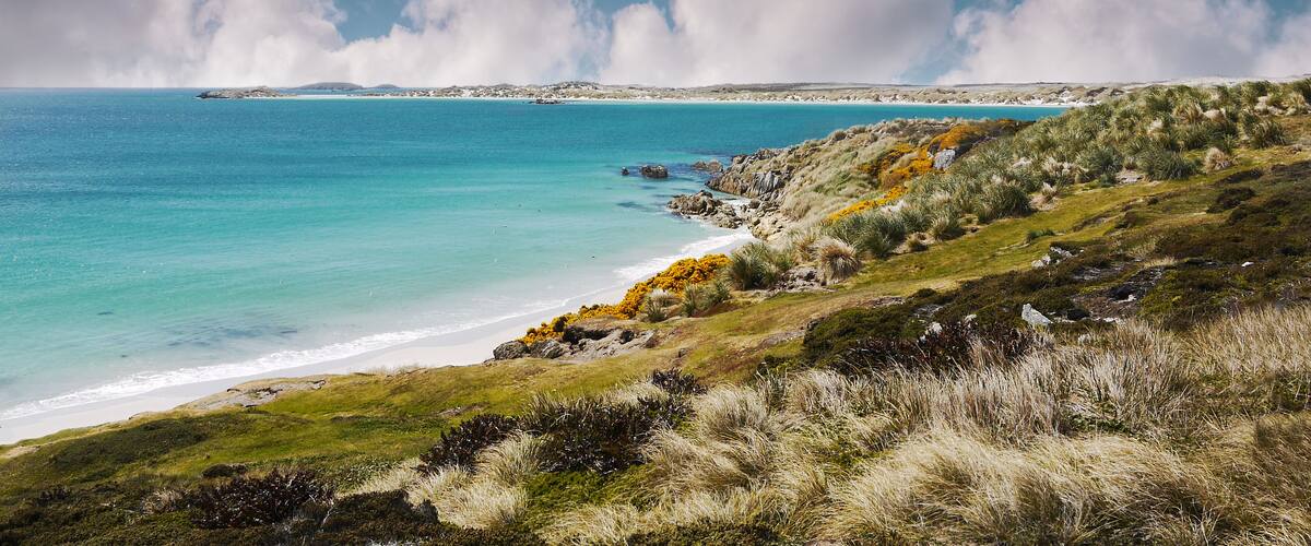 A coastal landscape with a clear blue sea, sandy beach, grassy hills, and rocky outcrops.
