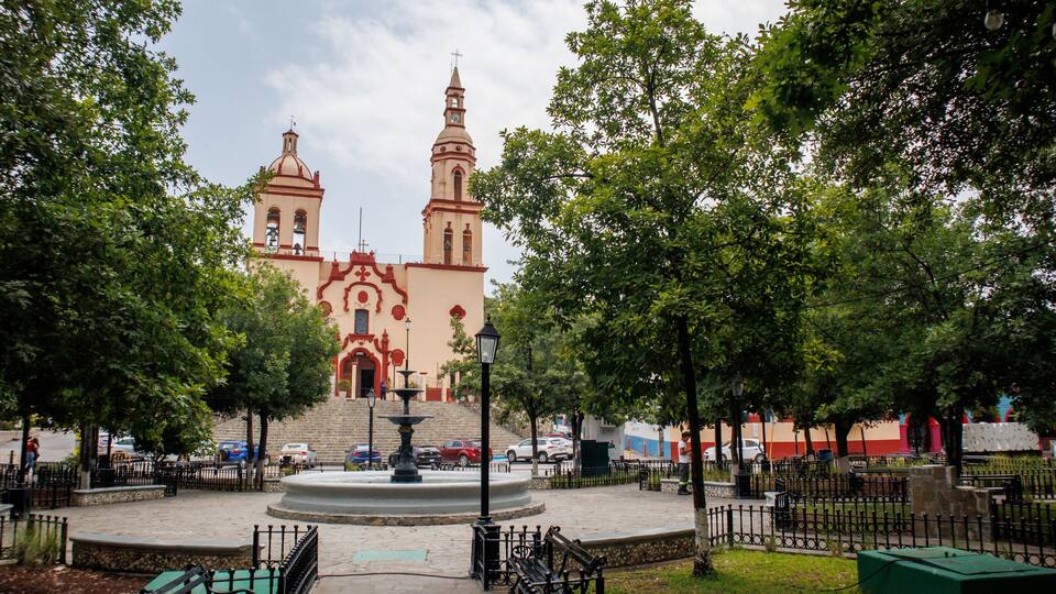 A church with two towers, a fountain, and surrounding trees.