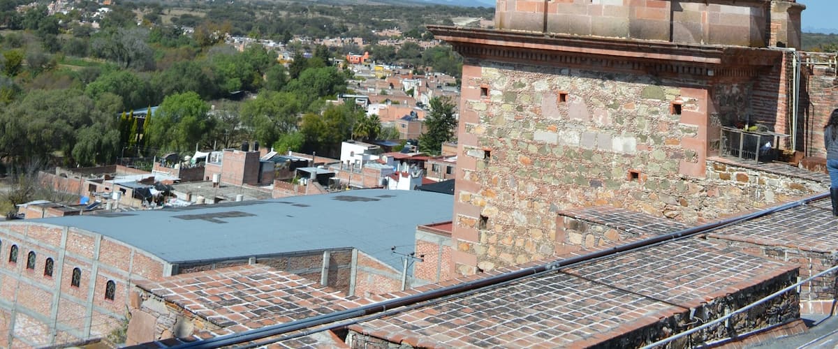 A historic bell tower with a cross on top, surrounded by a hilly landscape and a clear blue sky.