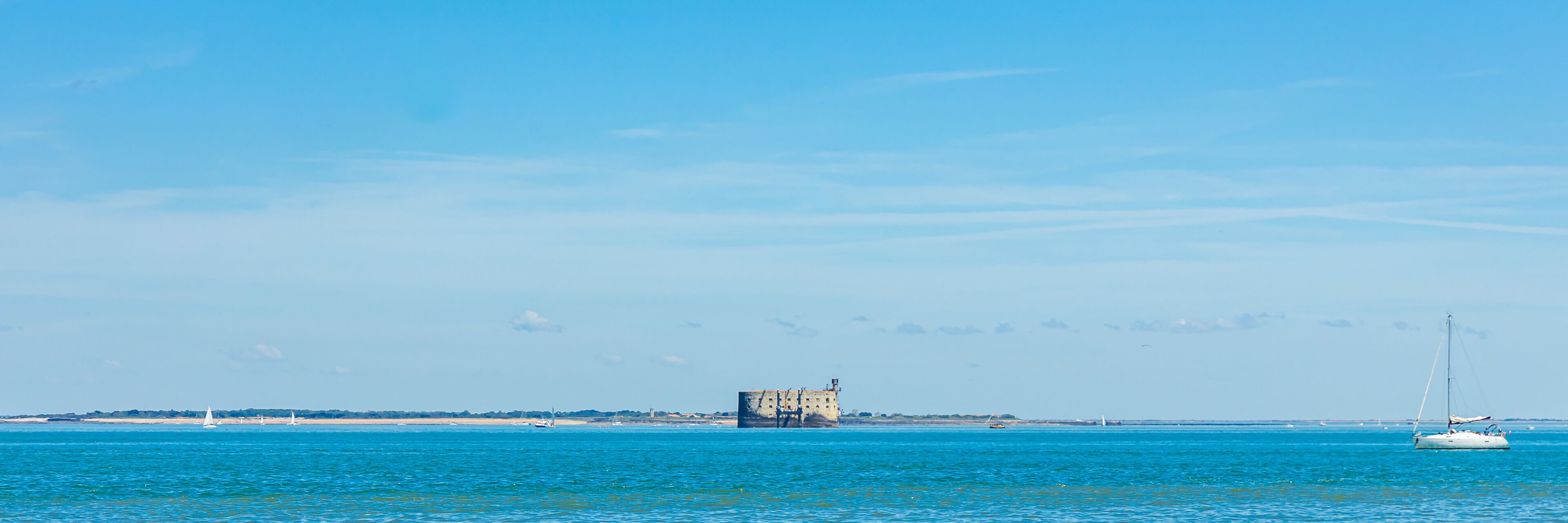 A coastal scene with a historic building, a sailboat, and a clear blue sky.