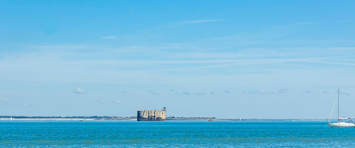 A coastal scene with a historic building, a sailboat, and a clear blue sky.
