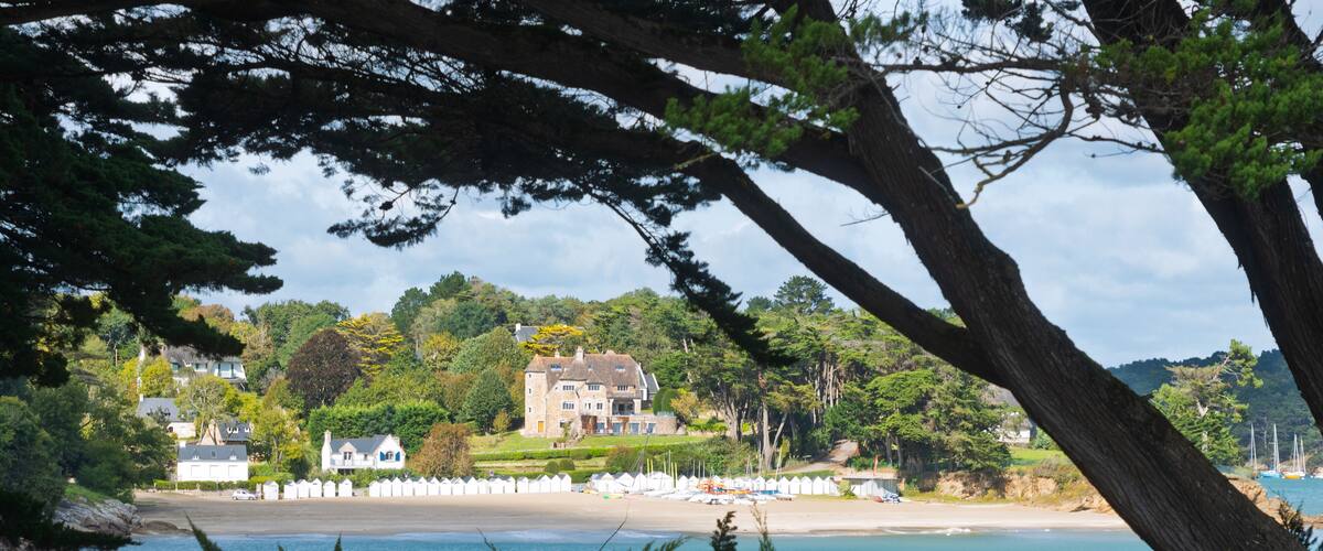 A coastal landscape with a large house surrounded by lush greenery and a beach with boats.
