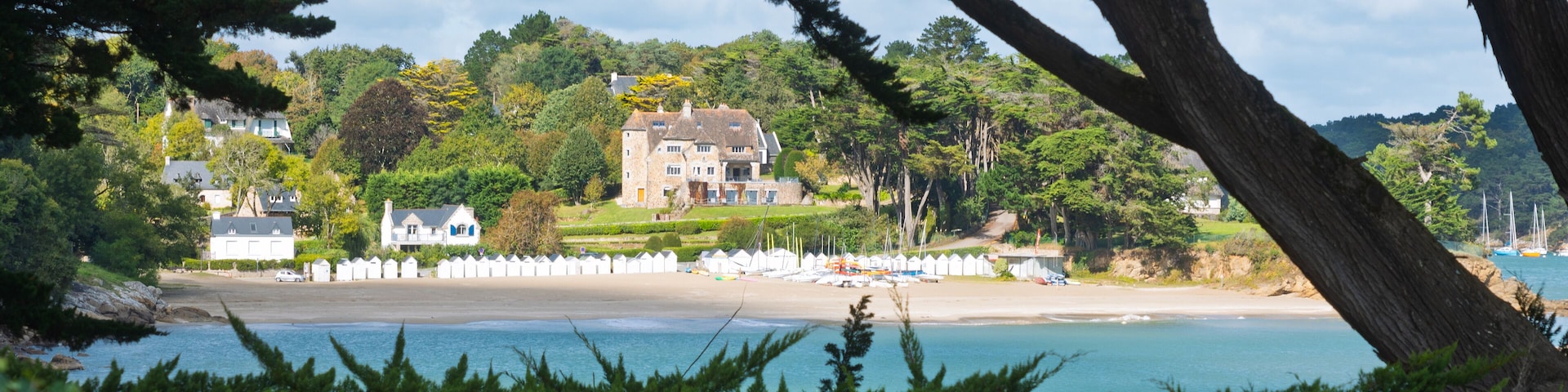 A coastal landscape with a large house surrounded by lush greenery and a beach with boats.