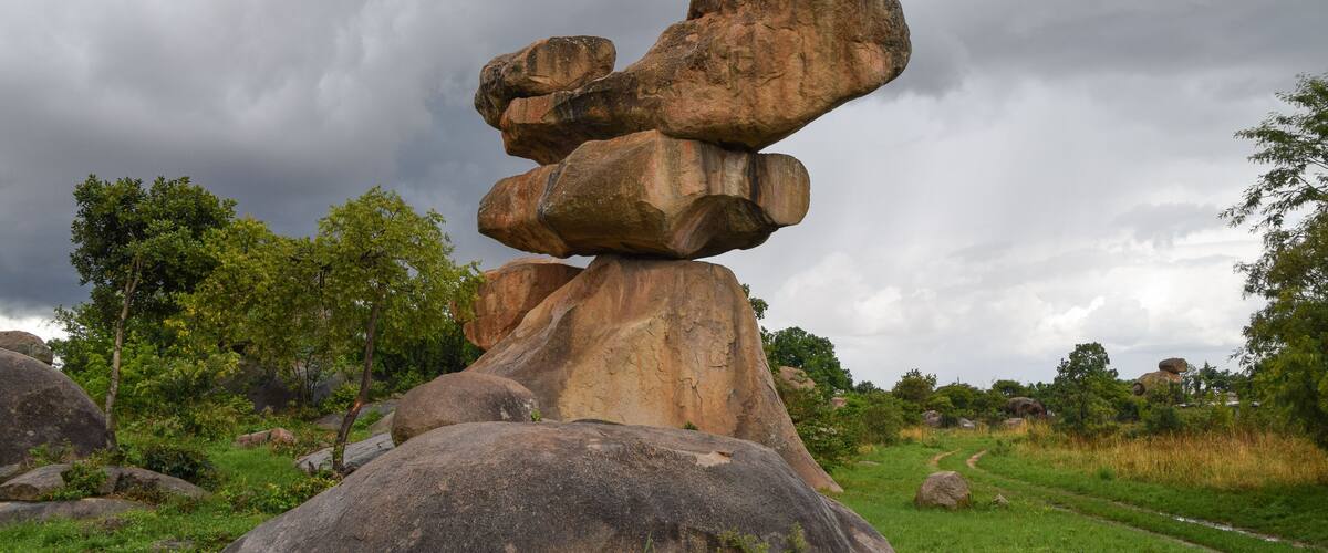 A large balanced rock formation in a grassy field with trees and a cloudy sky.
