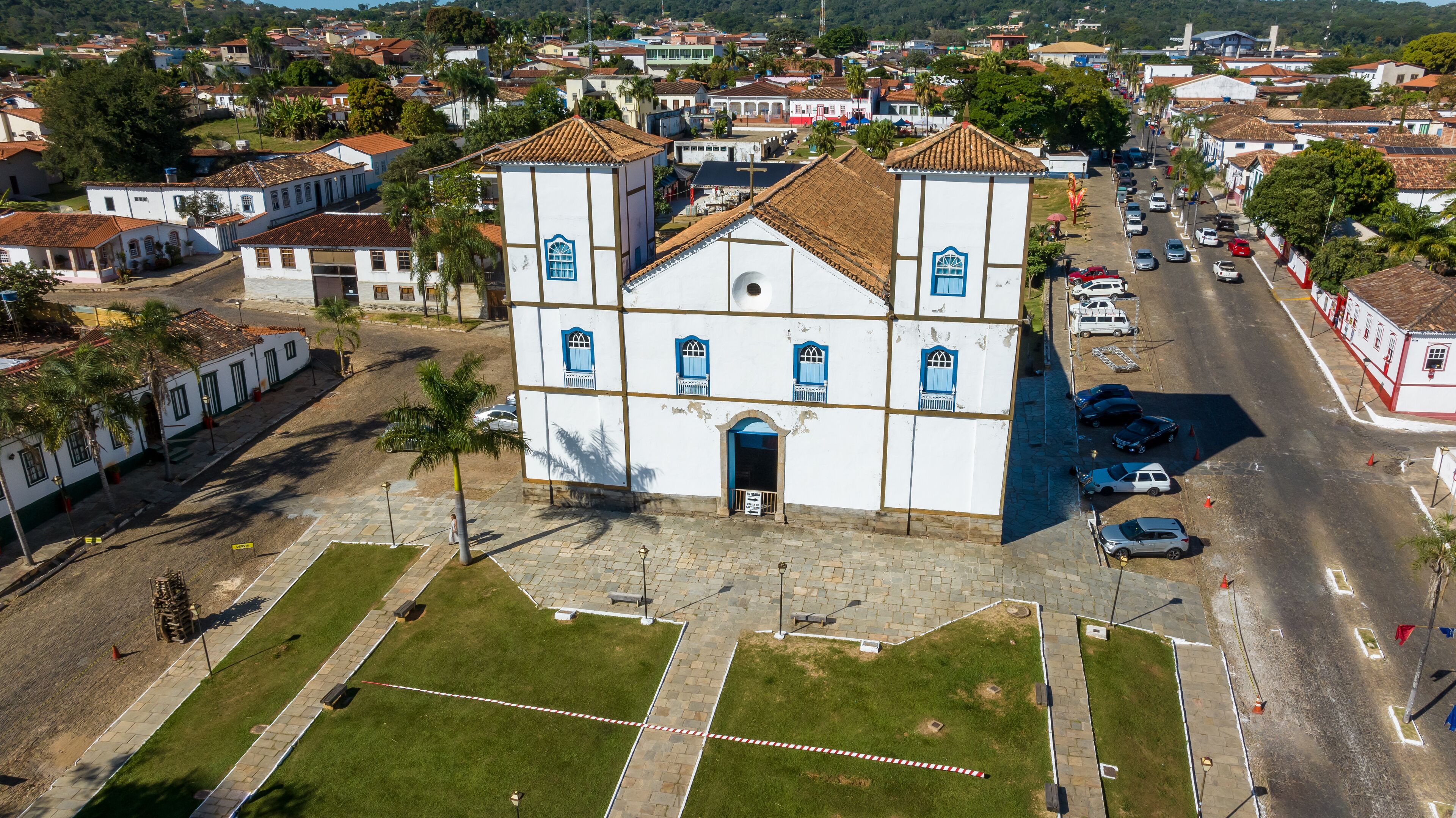 An aerial view of a town with a prominent white church with blue windows and a tiled roof, surrounded by residential buildings and a main road.