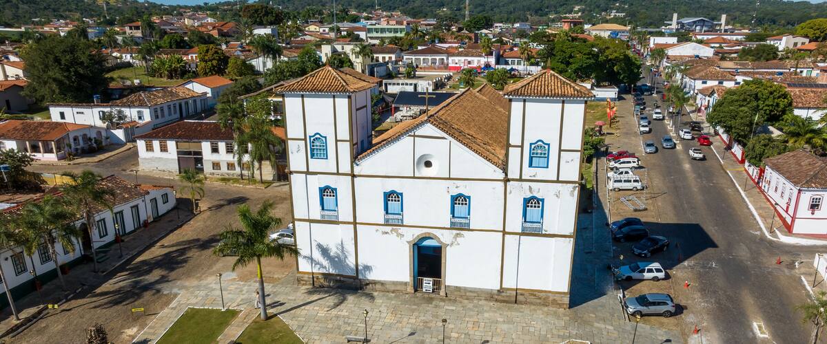 An aerial view of a town with a prominent white church with blue windows and a tiled roof, surrounded by residential buildings and a main road.