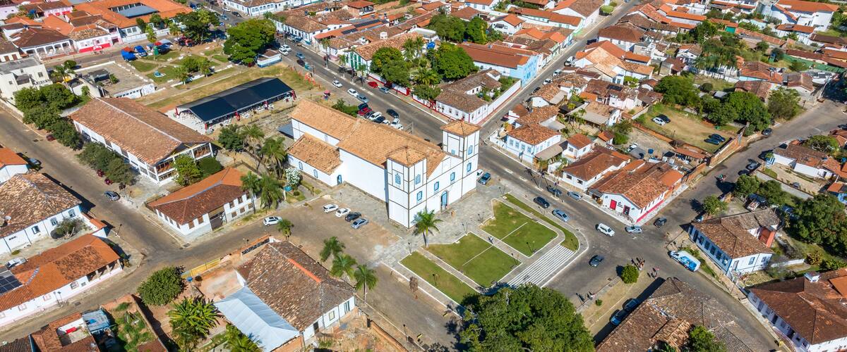 An aerial view of a residential area with red-tiled roofs, white buildings, and a central plaza with a large open space and a few trees.