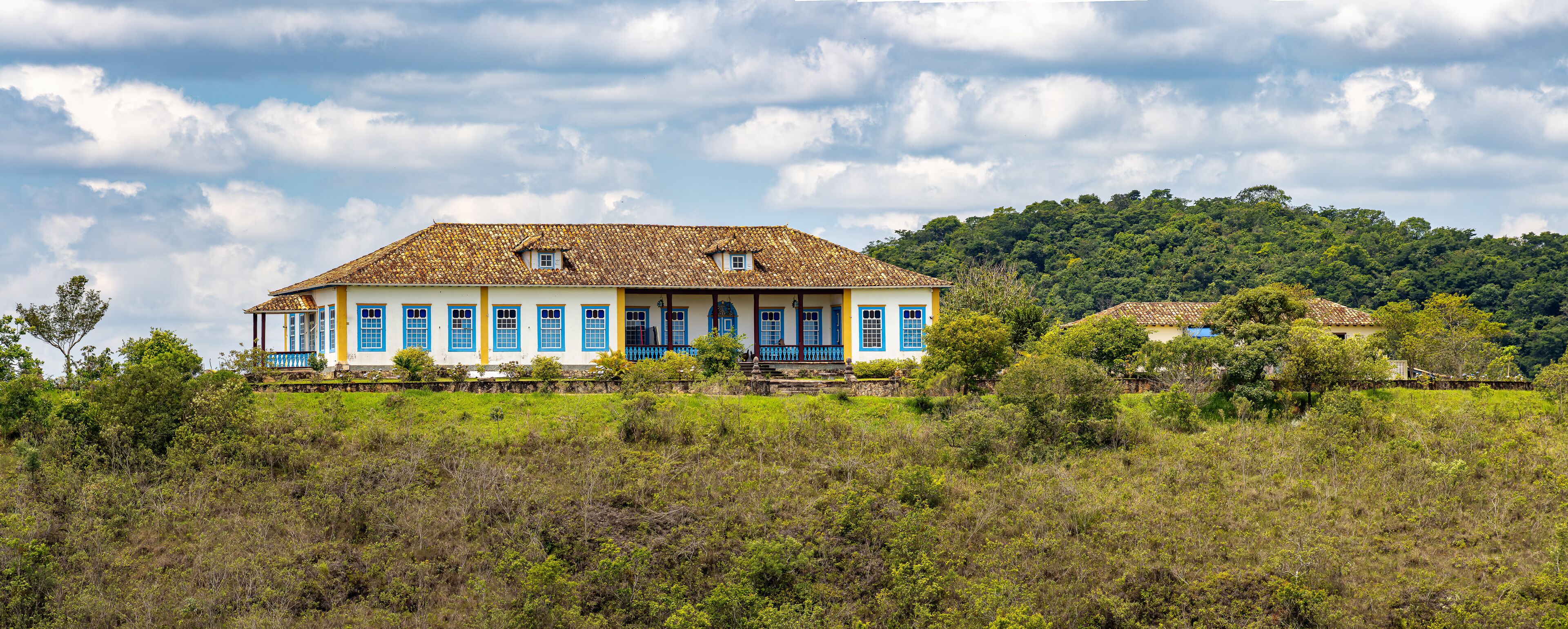 A large colonial-style house with blue doors and windows, surrounded by lush greenery and a hill in the background.