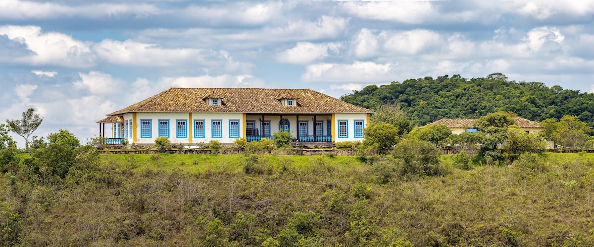 A large colonial-style house with blue doors and windows, surrounded by lush greenery and a hill in the background.
