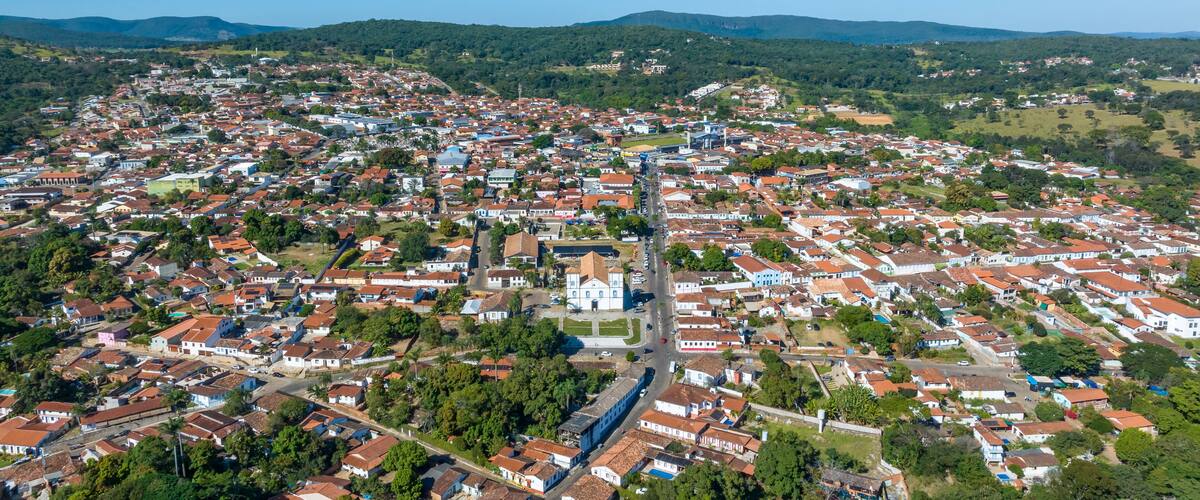 An aerial view of a densely populated town with red-roofed houses, surrounded by green hills and a clear blue sky.