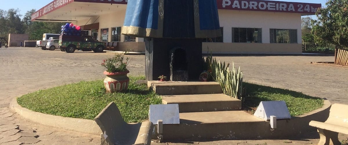 A statue of a religious figure with a blue robe and a red crown, located in front of a building with a sign that reads "CHU PADROEIRA 24h".