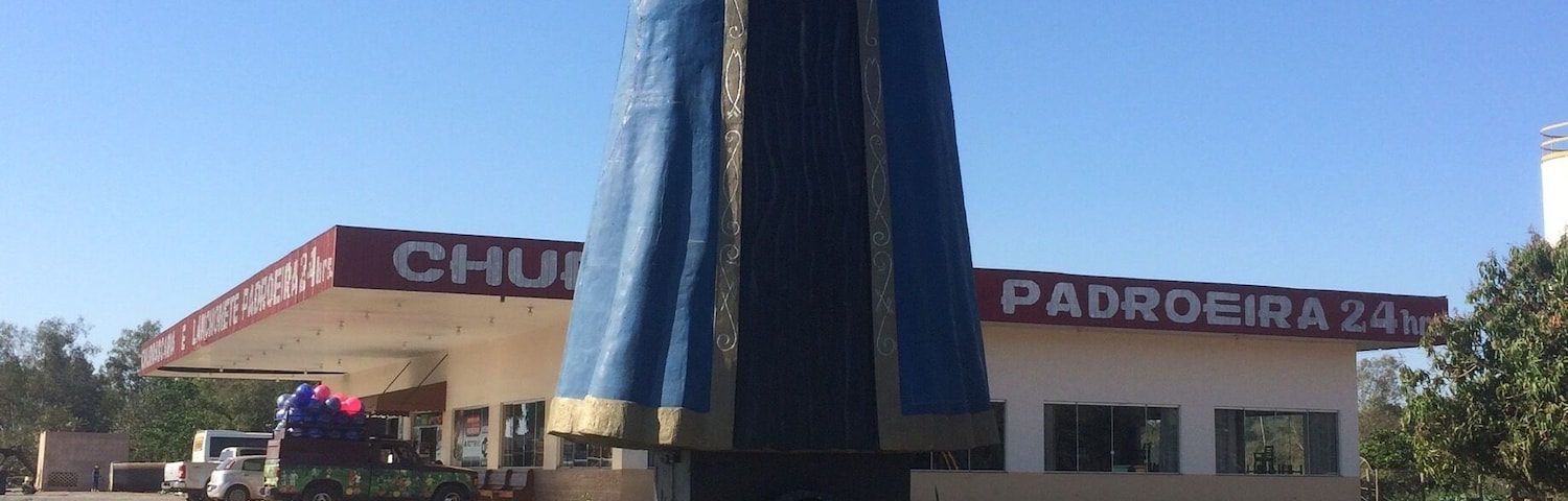 A statue of a religious figure with a blue robe and a red crown, located in front of a building with a sign that reads "CHU PADROEIRA 24h".