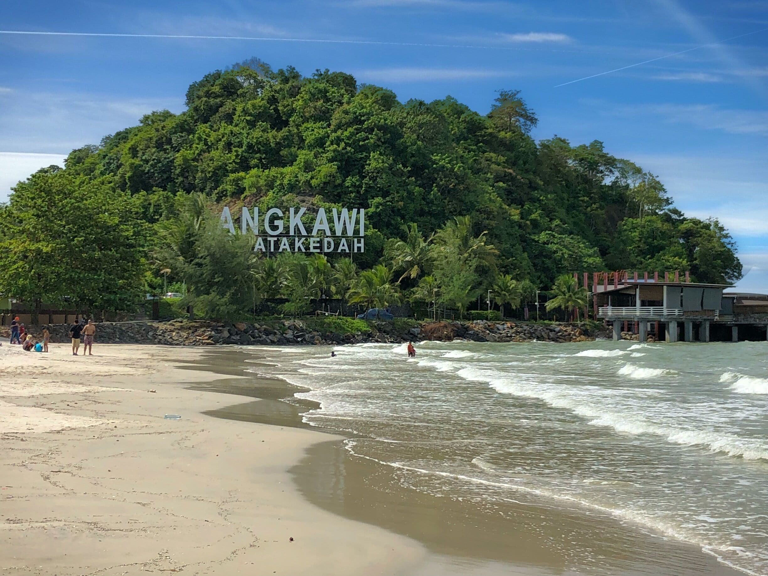A beach with waves, a forested hill, and a building with 'ANGKAWI ATAKEDAH' written on it.