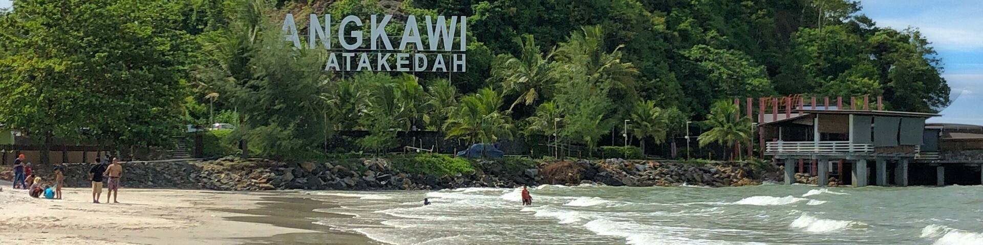 Una playa con olas, una colina boscosa y un edificio con la inscripción 'ANGKAWI ATAKEDAH'.
