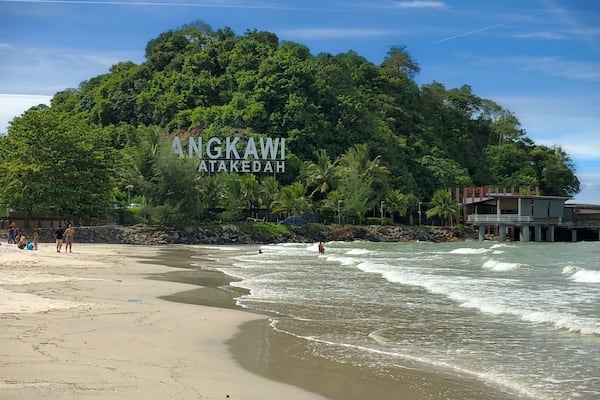 Une plage avec des vagues, une colline boisée et un bâtiment sur lequel on peut lire l’inscription « ANGKAWI ATAKEDAH ».