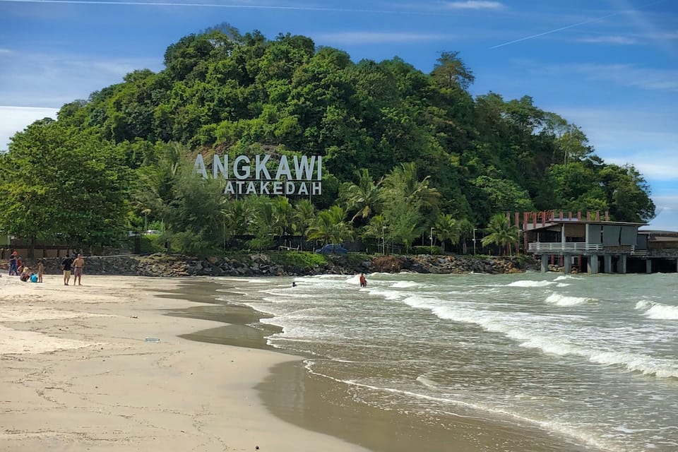 A beach with waves, a forested hill, and a building with 'ANGKAWI ATAKEDAH' written on it.