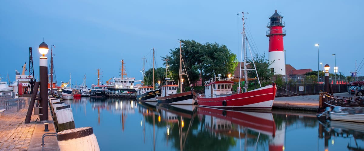 A calm harbor with a prominent red and white lighthouse, several moored boats, and a clear reflection in the water.
