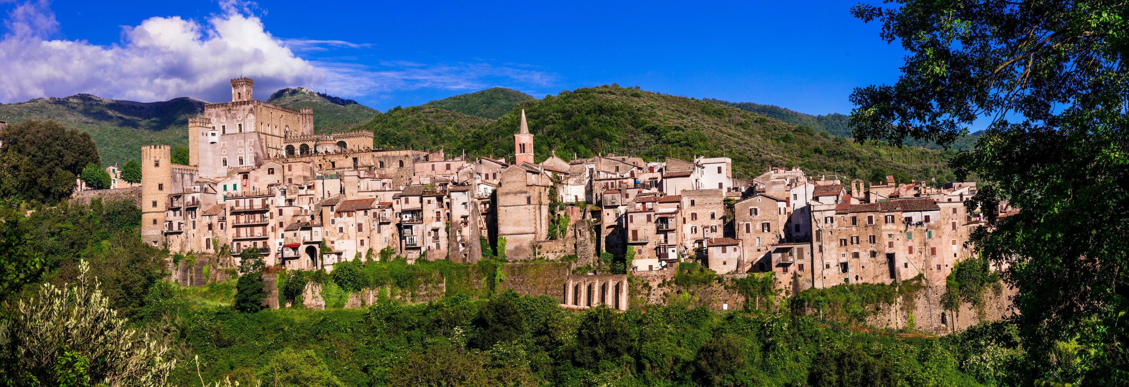A historic town with medieval architecture, a prominent tower, and lush green hills in the background.