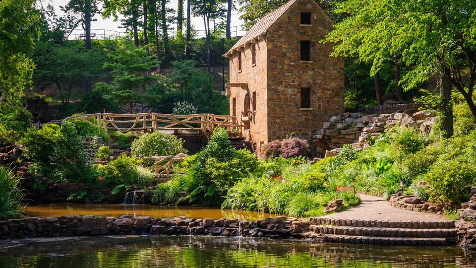 A stone building with a wooden bridge over a pond, surrounded by lush greenery and trees.