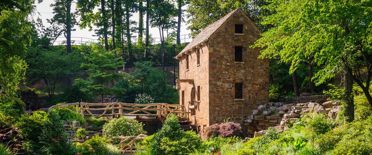 A stone building with a wooden bridge over a pond, surrounded by lush greenery and trees.