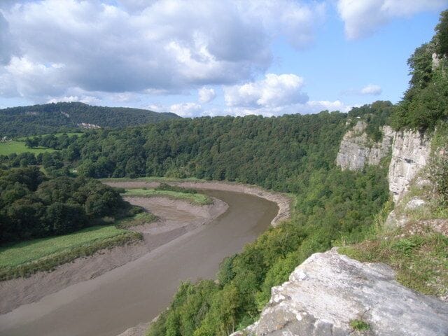 A river winding through a green valley with a forested hillside and a rocky cliff.