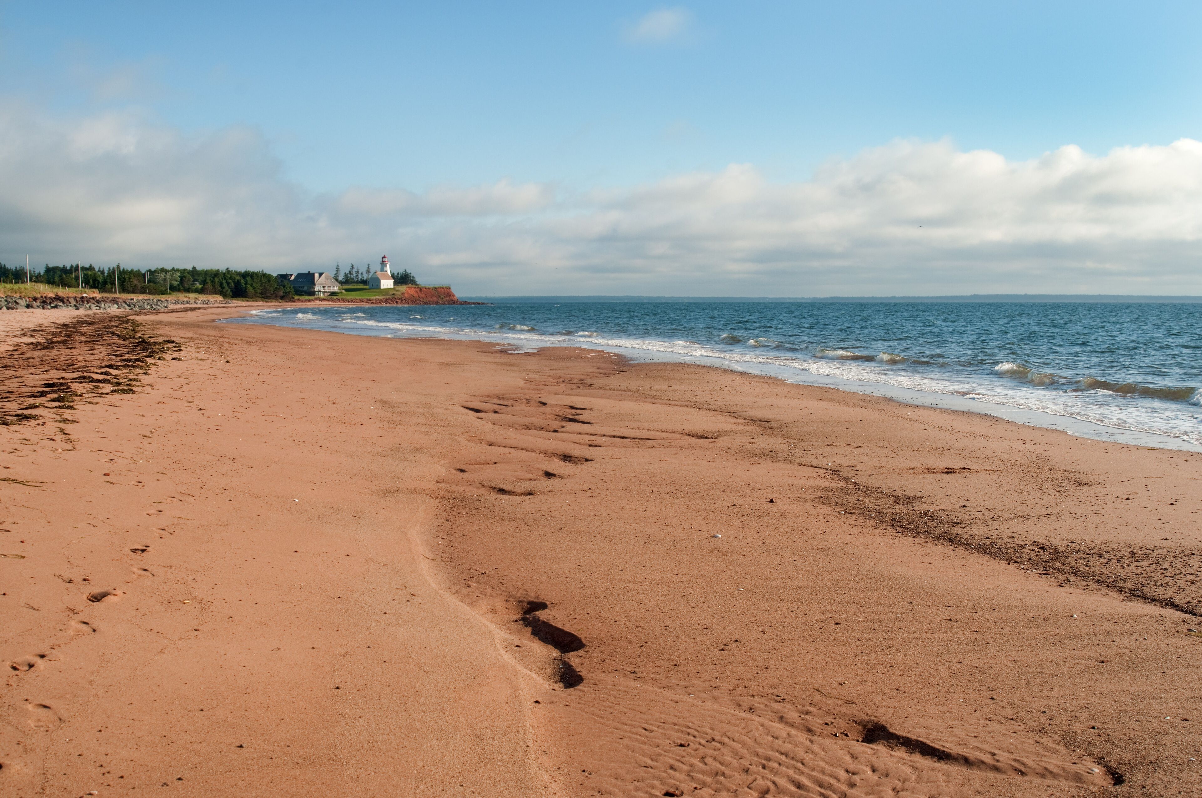 A sandy beach with footprints leading to the water, a lighthouse in the distance, and a clear sky.