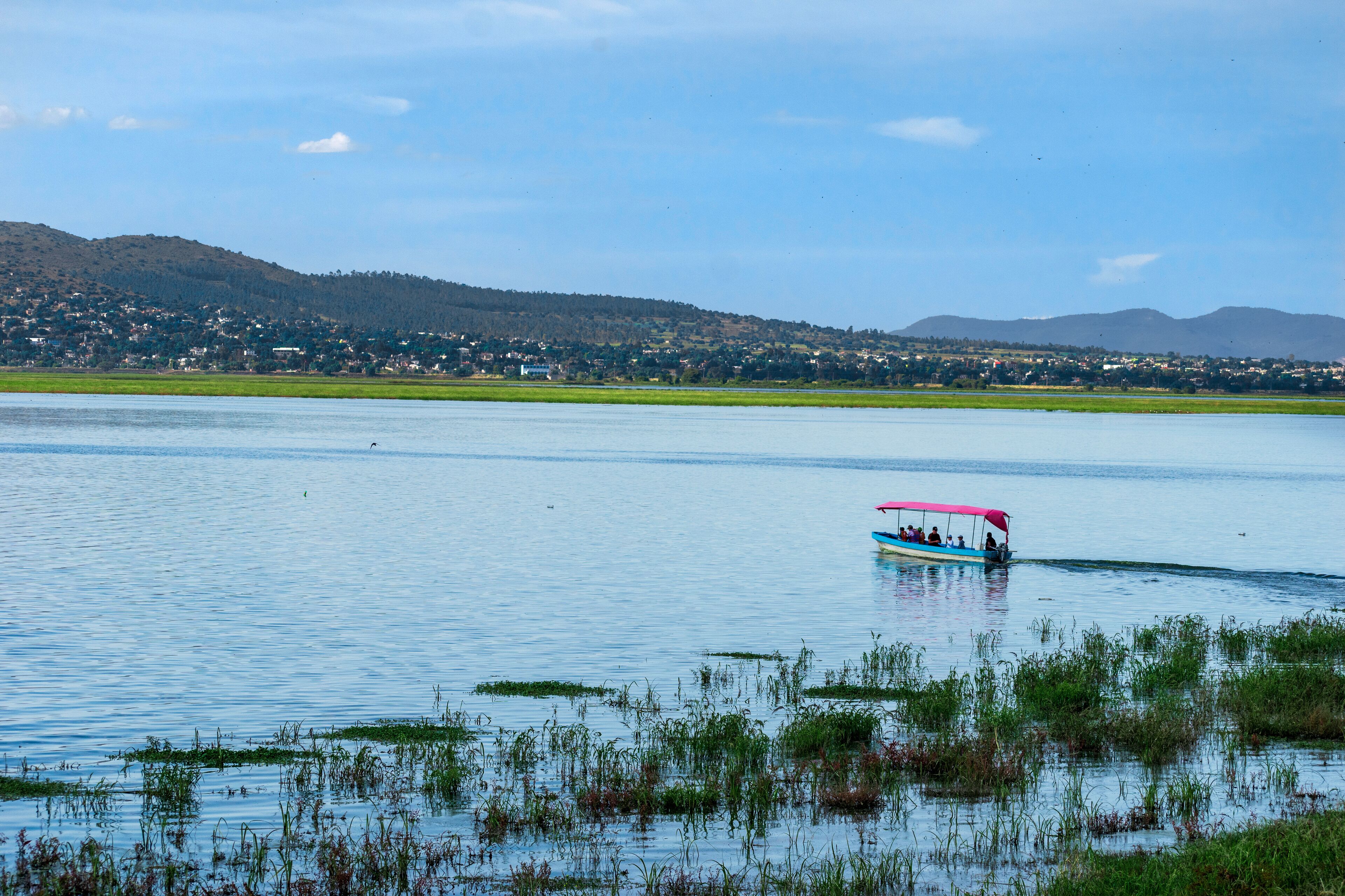 A lake with a boat carrying passengers, surrounded by greenery and distant hills.