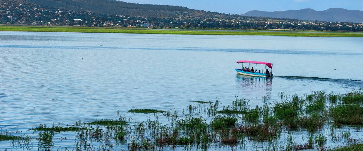 A lake with a boat carrying passengers, surrounded by greenery and distant hills.