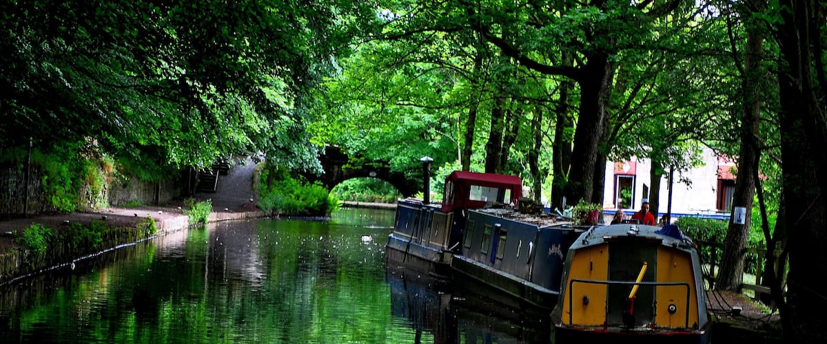 A narrow canal with a narrowboat docked on the right, surrounded by lush green trees and a stone bridge in the background.