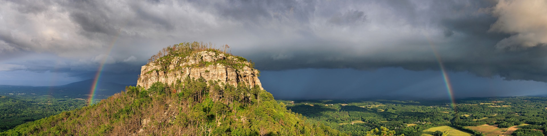 Montanha com pico arborizado, céu tempestuoso e arco-íris.