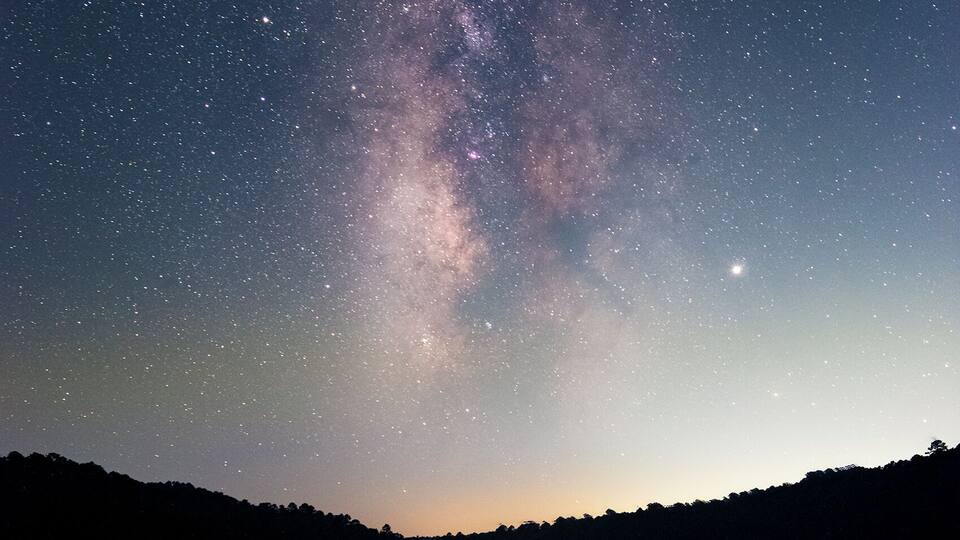 A starry night sky over a calm lake with a silhouette of trees in the foreground.