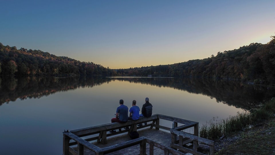 A calm lake with three people sitting on a wooden dock, surrounded by autumn-colored trees.