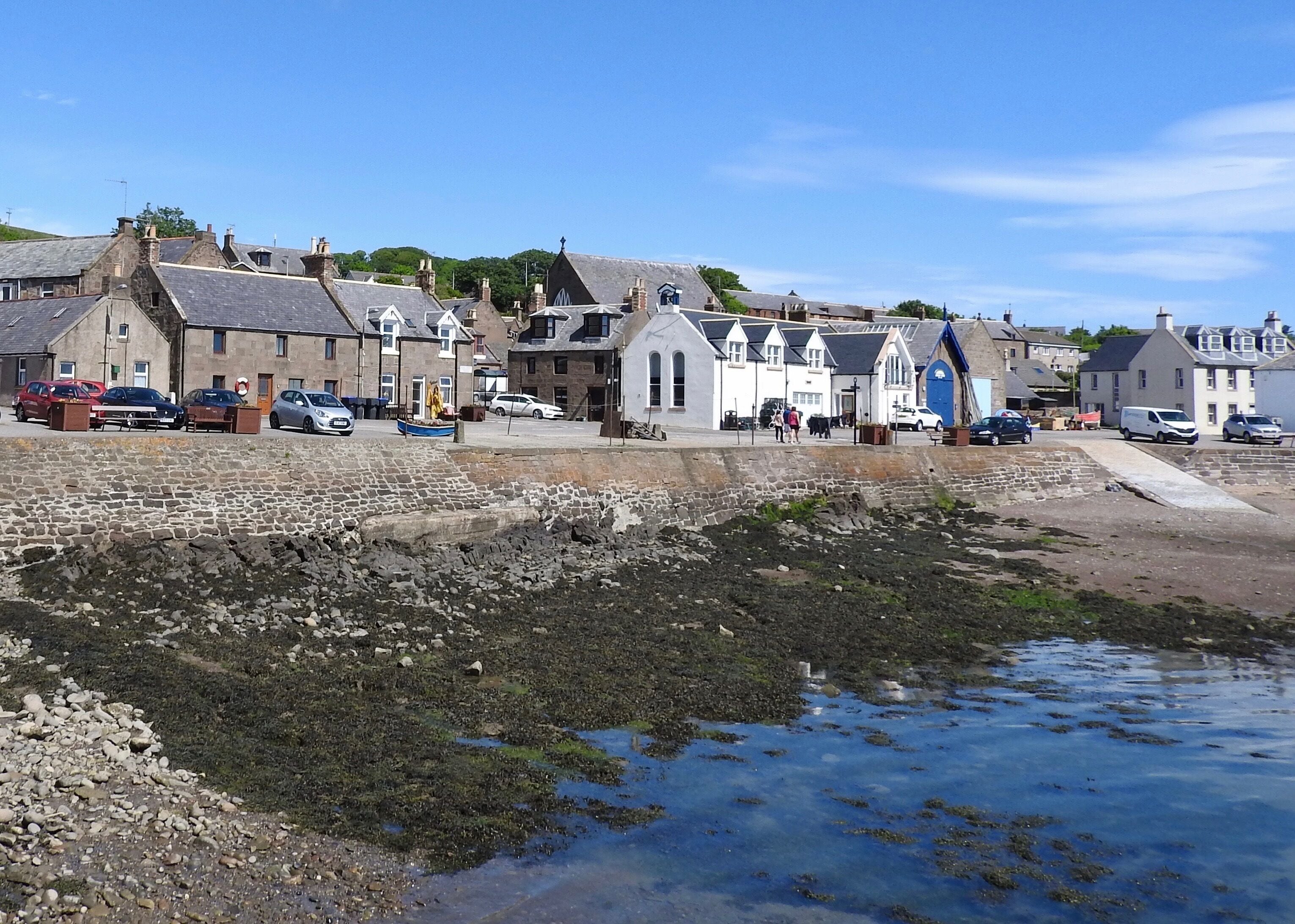 A coastal town with stone buildings, parked cars, and a rocky shoreline.