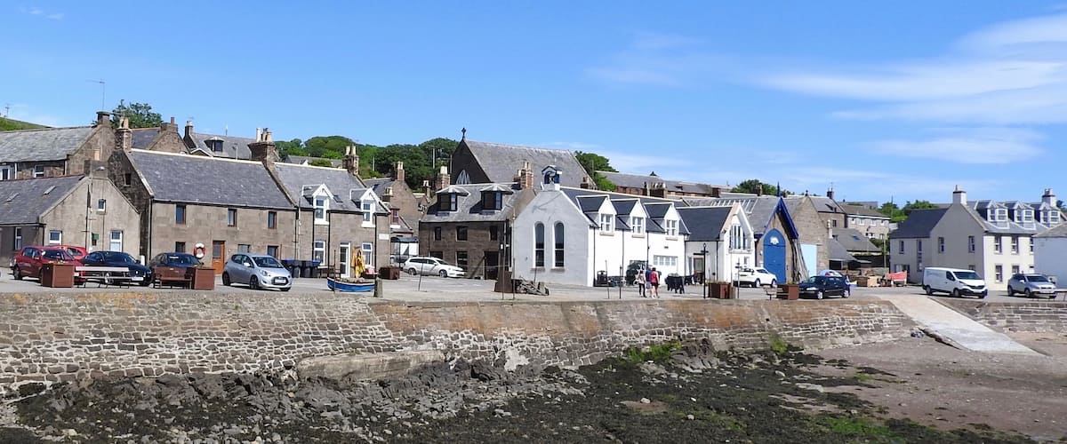 A coastal town with stone buildings, parked cars, and a rocky shoreline.