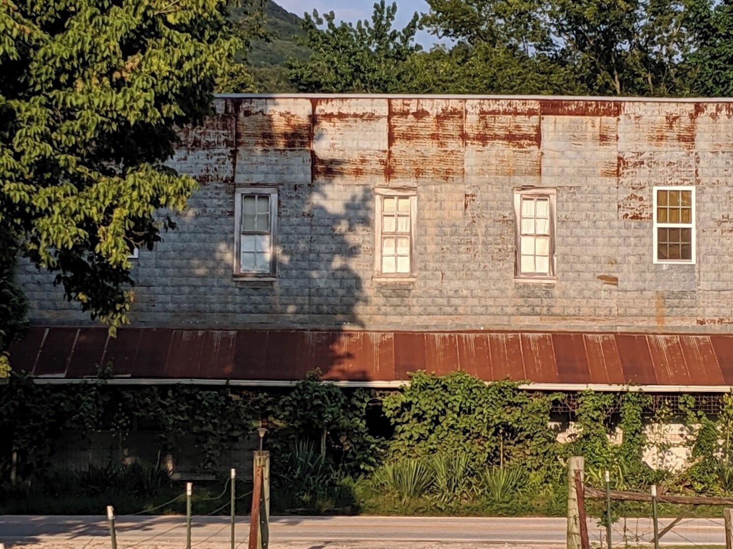 A weathered, rusted building with multiple windows and a metal roof, surrounded by greenery and trees.