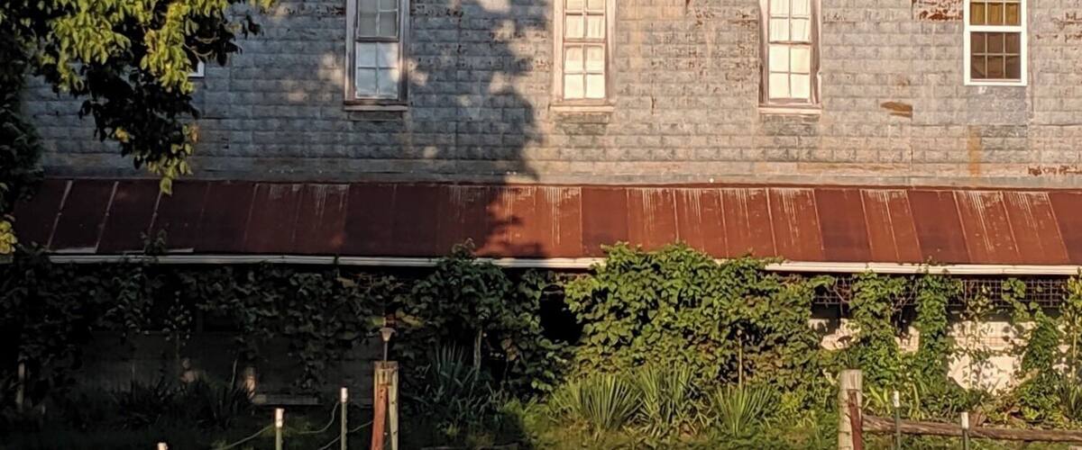 A weathered, rusted building with multiple windows and a metal roof, surrounded by greenery and trees.