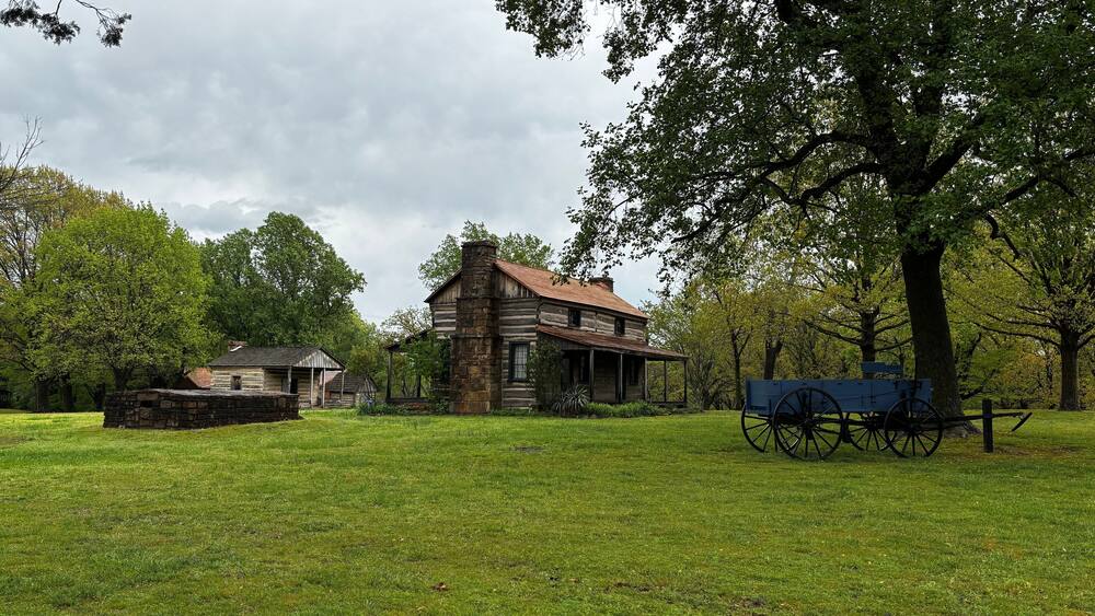 A historic log cabin with a covered porch, surrounded by lush green grass and trees.