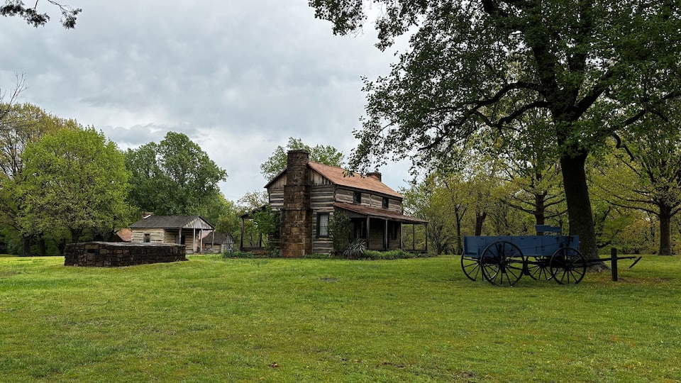 A historic log cabin with a covered porch, surrounded by lush green grass and trees.