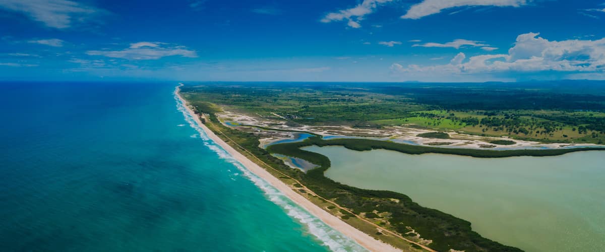 An aerial view of a coastal area with a long beach, green land, and a large body of water.
