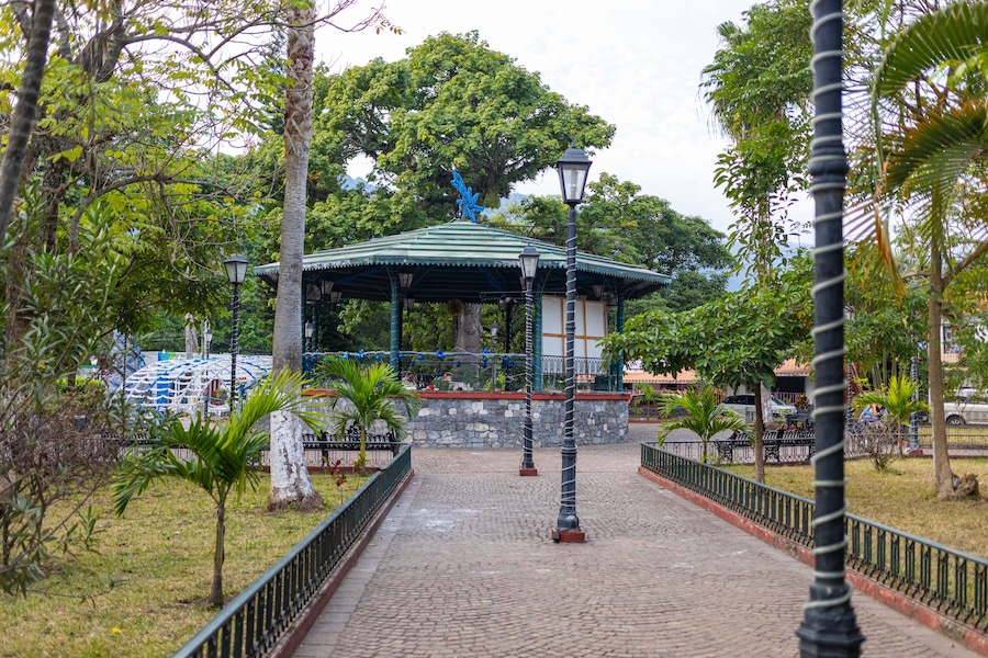 Un jardín con un pabellón de techo verde y detalles azules, rodeado de árboles y un camino adoquinado.