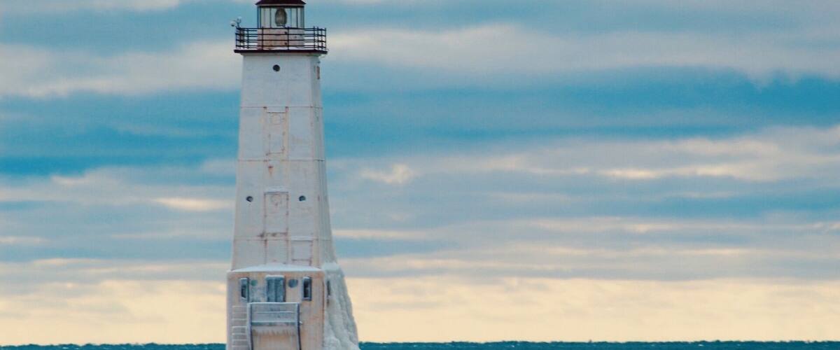 A tall, white lighthouse with a red top, surrounded by icy waters and a cloudy sky.