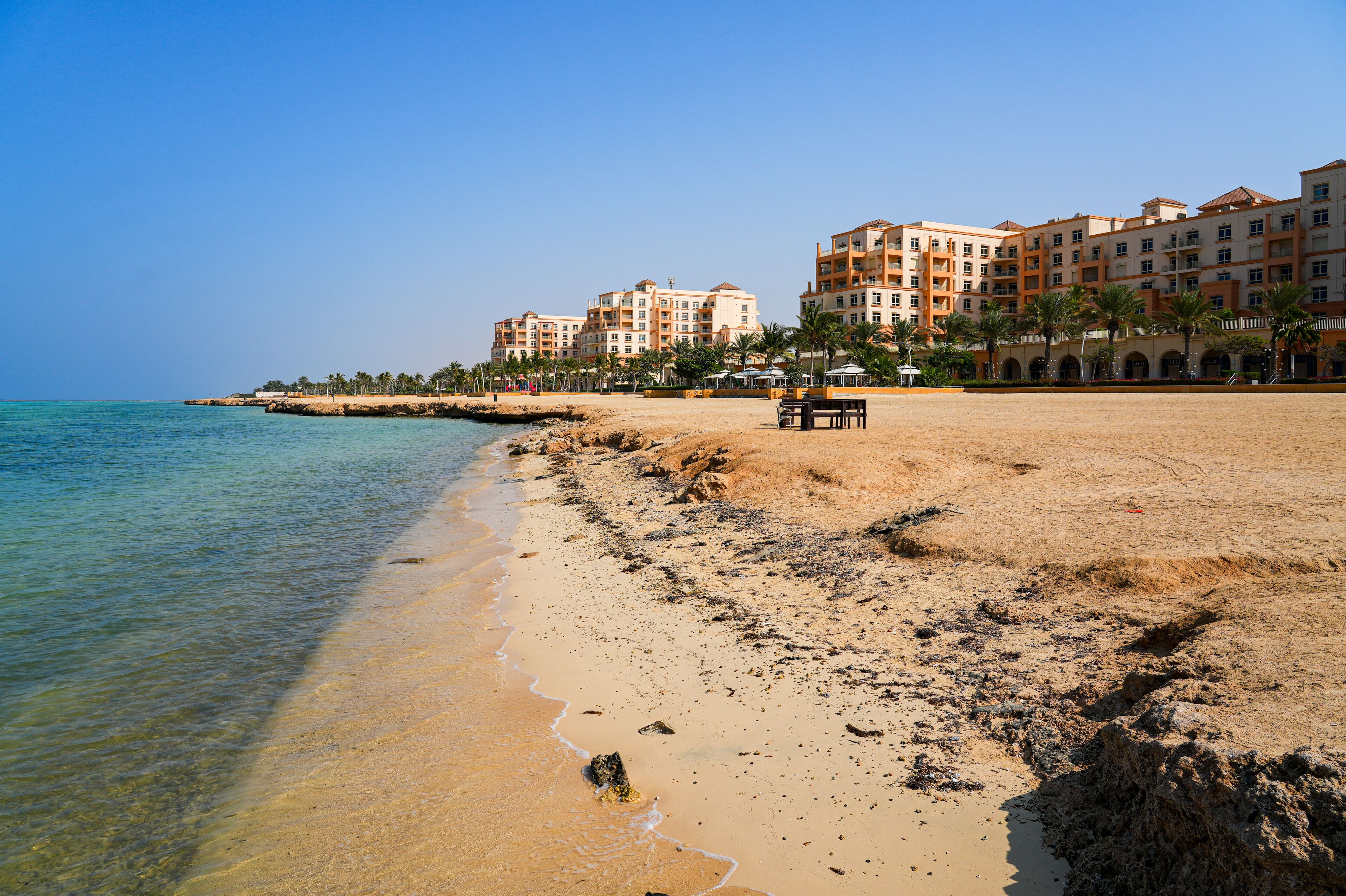 A sandy beach with clear water, palm trees, and a hotel in the background.